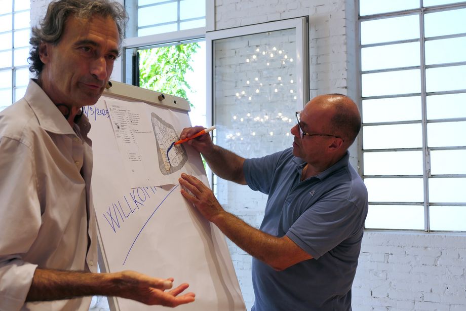 Zwei Männer stehen vor einer Flipchart in einem hellen Raum, ein Mann arbeitet an der Flipchart, der andere Mann spricht.