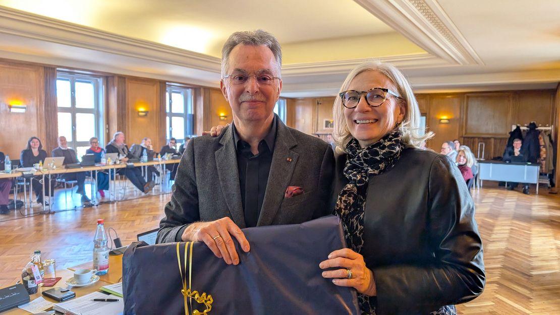 Ein Mann und eine Frau lächeln in die Kamera, im Hintergrund sitzen Personen auf Stühlen an einem Tisch.