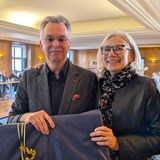 Ein Mann und eine Frau lächeln in die Kamera, im Hintergrund sitzen Personen auf Stühlen an einem Tisch.