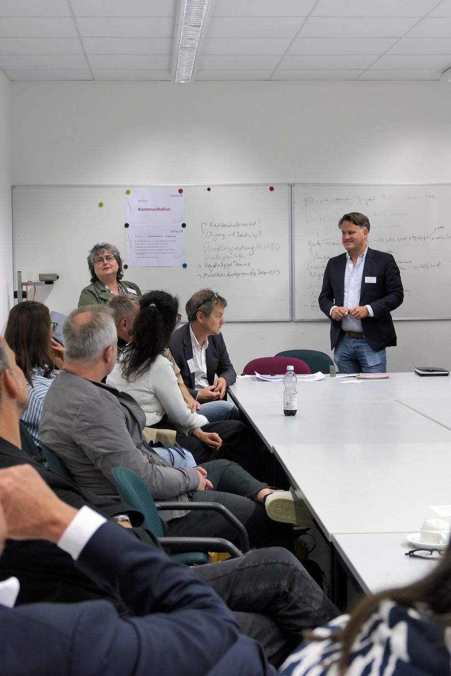 Eine Gruppe von Menschen sitzt an einem großen hellen Tisch. Sie blicken zu zwei Personen die vor einer hellen Wand stehen.