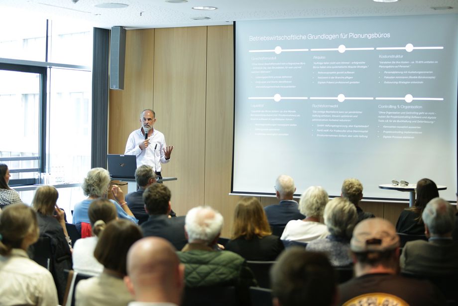 Ein Mann steht vor einer Leinwand, auf der eine Präsentation gezeigt wird, in der Hand hält er ein Mikrofon. Vor ihm sitzt ein Publikum, das ihm zuhört. - Foto: Ingo Lammert/Architektenkammer NRW