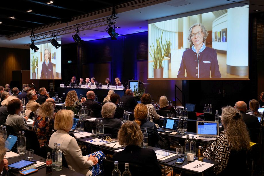 Viele Leute sitzen in einem Raum auf Stühlen, auf einer Leinwand wird ein Videogrußwort von  Ina Scharrenbach, Ministerin für Heimat, Kommunales, Bau und Digitalisierung des Landes NRW, gezeigt.