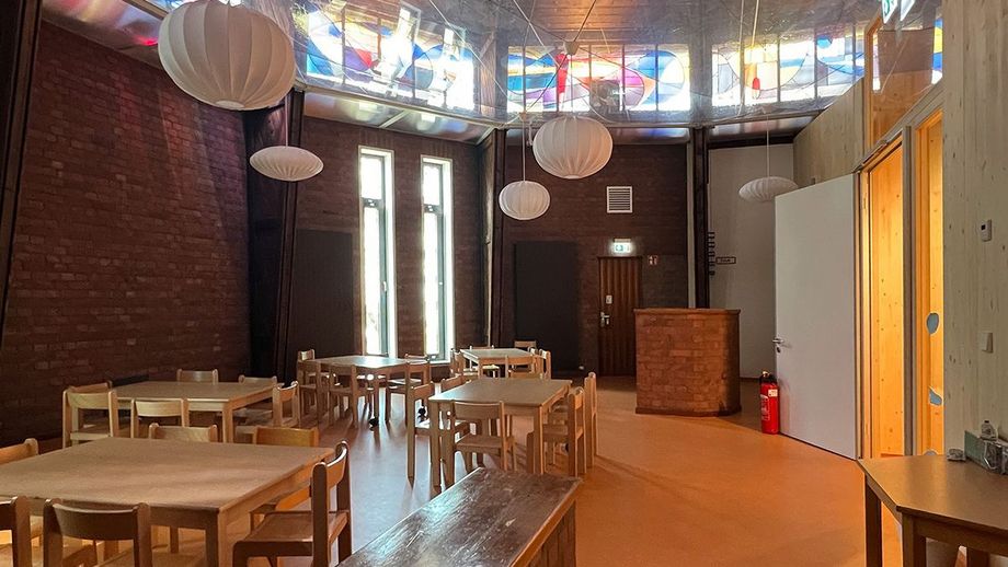 Blick in den ehemaligen Altarraum mit Holztischen und Stühlen für die Kinder, weißen Pendellampen. Oben im Bild die ehemaligen bunten Kirchenfenster