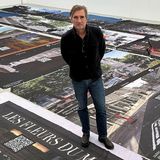 Ein Mann steht in einem Raum und lächelt in die Kamera, Banner liegen auf dem Boden und zeigen Kunstprojekte.