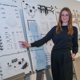 Eine Frau steht vor einem Plakat und lächelt in die Kamera.