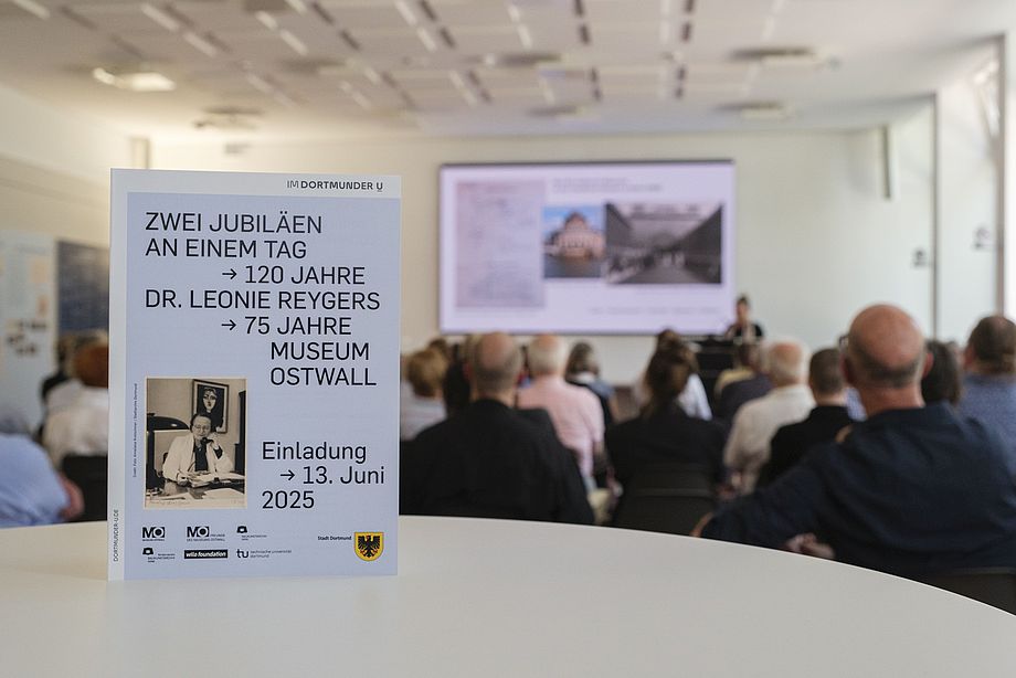 Rund 50 Teilnehmer*innen nahmen an der Veranstaltung im Gartensaal des Baukunstarchivs NRW teil. - Foto: Detlef Podehl - Das Bild zeigt einen Ausschnitt des Gartensaals des Baukunstarchivs NRW, im Vordergrund ist ein Einladungsflyer zur Veranstaltung "120 Jahre Dr. Leonie Reygers / 75 Jahre Museum Ostwall", im Hintergrund sitzen Personen, die einem Vortrag zuhören.