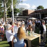 Das Foto zeigt viele Menschen auf einem begrünten Platz, die sich in Kleingruppen unterhalten bei sommerlicher Atmosphäre.