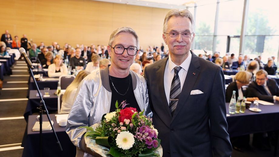 Eine Frau mit Blumenstrauß und ein Mann vor einem mit Publikum besetzten Saal