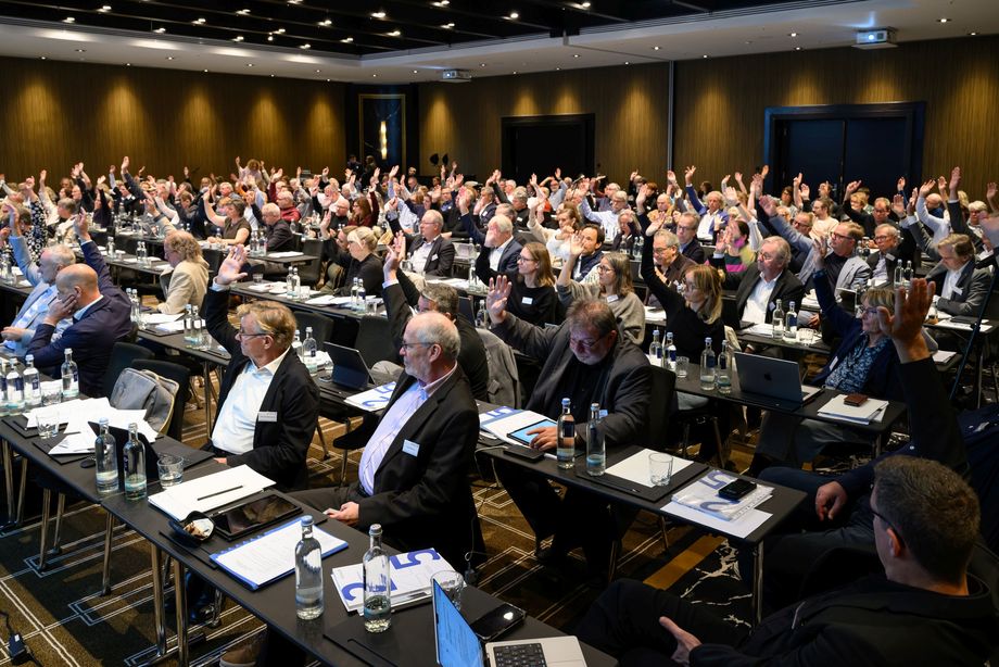 Zahlreiche Menschen sitzen in einem Raum, einige von ihnen heben die Hand zu einem Handzeichen.