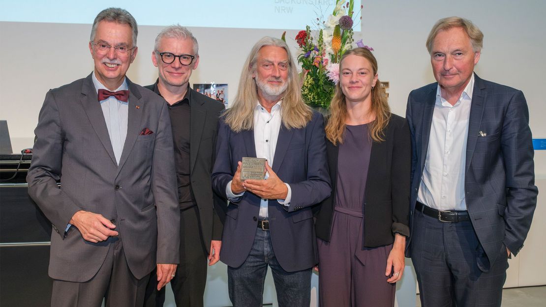 Eine Gruppe Menschen schaut und lächelt in eine Kamera. Ein Mann in der Mitte zeigt eine Medaille, u.a. mit der Aufschrift "Friedrich Halstenberg Medaille". - Foto: Detlef Podehl