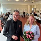 Zwei in die Kamera lächelnde Personen vor dem Hintergrund der Tischrunde