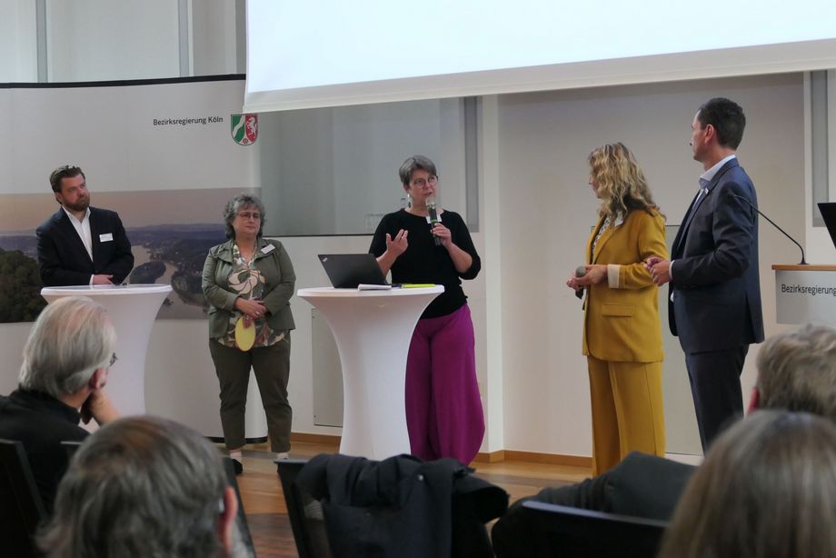 Eine Gruppe von Frauen und Männern steht vor sitzendem Publikum und unterhält sich.