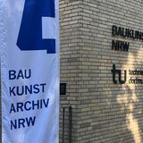 Der Eingangsbereich des Gebäudes "Baukunstarchiv NRW". An der Fassade hängen die Schriftzüge "Baukunstarchiv NRW" und "Technische Universität Dortmund". Eine Beachflag in den Farben weiß und blau mit der Aufschrift "Baukunstarchiv NRW" steht im Vordergrund.
