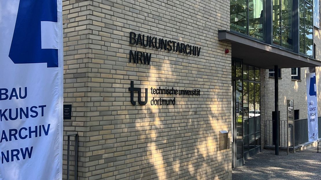 Baukunstarchiv NRW - Eingangsbereich Der Eingangsbereich des Gebäudes "Baukunstarchiv NRW". An der Fassade hängen die Schriftzüge "Baukunstarchiv NRW" und "Technische Universität Dortmund". Eine Beachflag in den Farben weiß und blau mit der Aufschrift "Baukunstarchiv NRW" steht im Vordergrund.