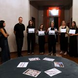 Eine Gruppe junger Personen steht in einem Raum beisammen, teilweise halten sie Plakate. Vor ihnen steht ein Tisch, auf dem Plakate liegen.