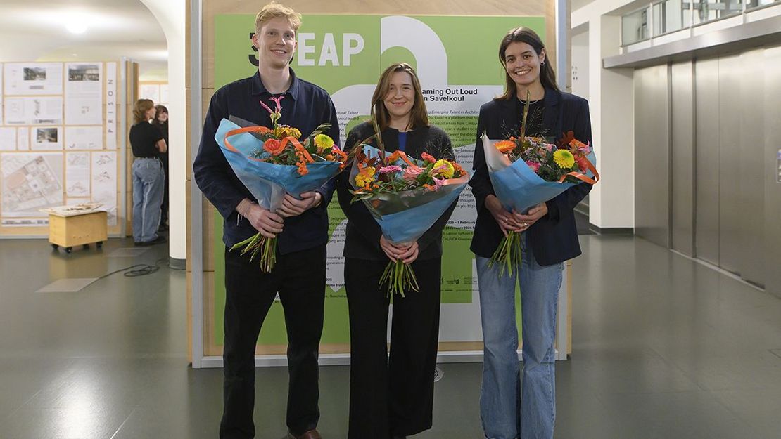 Zwei Frauen und ein Mann mit Blumensträußen in der Hand vor einer Plakatwand