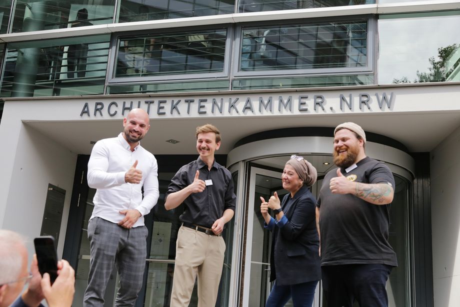 Eine Gruppe junger Leute steht vor einem Gebäude mit der Aufschrift "ARCHITEKTENKAMMER.NRW". Sie werden fotografiert und lächeln und zeigen freundliche Handgesten. - Foto: Ingo Lammert/Architektenkammer NRW