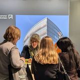 Vier Personen stehen an einem Infostand und unterhalten sich, im Hintergrund ist ein Banner mit einem Haus abgedruckt.