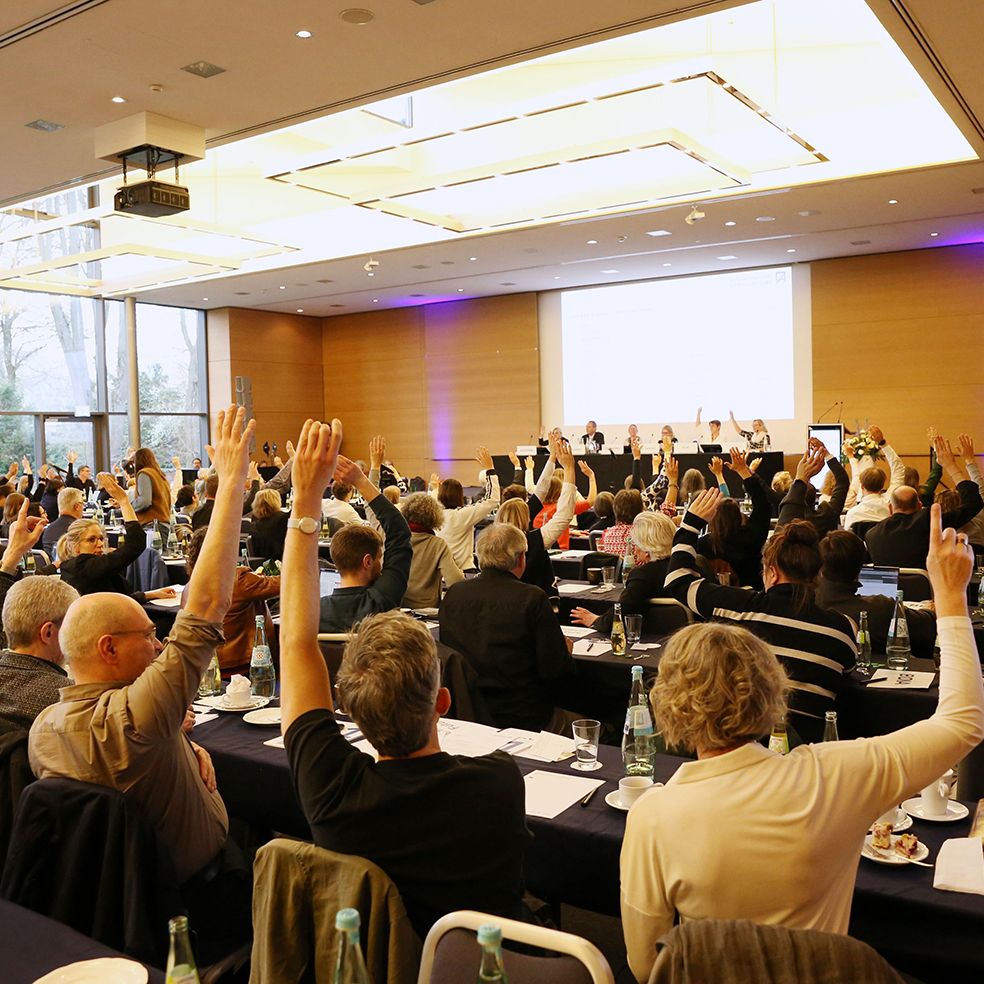 Konferenzsaal mit vielen Menschen, die zur Abstimmung die Hände erhoben haben, von hinten fotografiert.