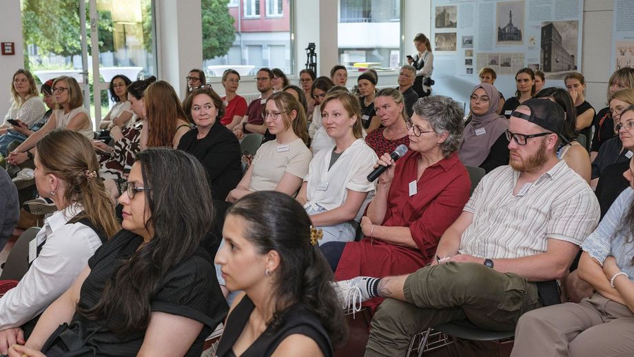 Impression: Aufnahme des voll besetzten Veranstaltungssaals im Baukunstarchiv. Männer und Frauen sitzen in Stuhlreihen und blicken nach vorne Richtung Vortragende. Eine Dame aus dem Plenum hält ein Mikro in der Hand und spricht.