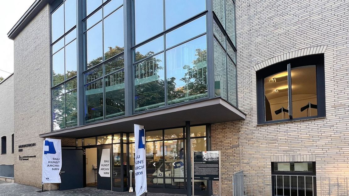 Baukunstarchiv NRW in Dortmund. Eine Hausfassade mit großer Fensterfront und kleinen Flaggen vor der Eingangstür.