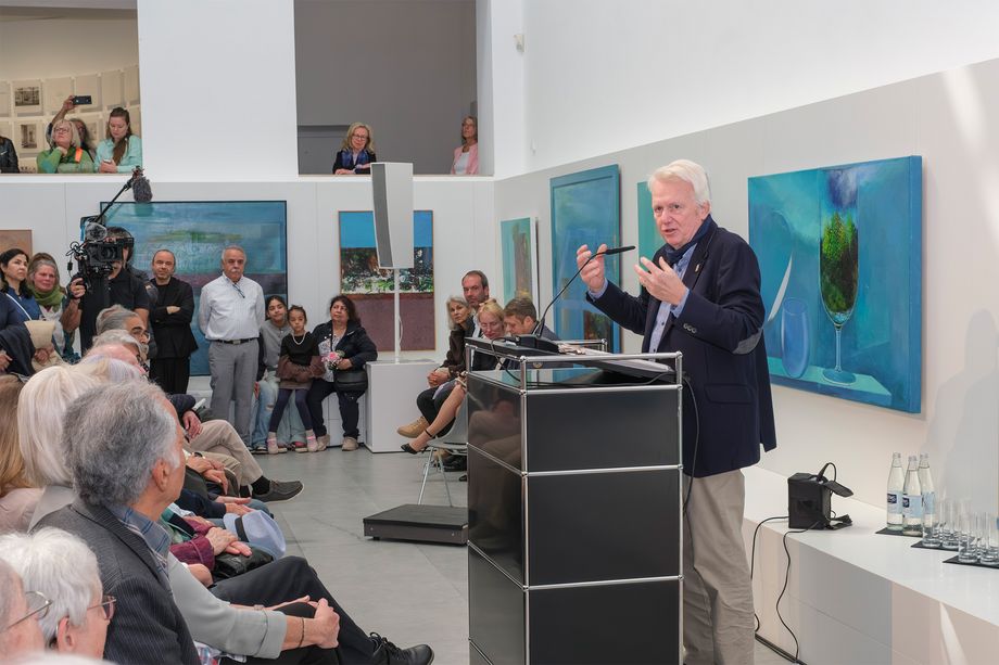 Menschen sitzen in einem Ausstellungsraum, an den Wänden hängen bunte Gemälde. Eine Person steht an einem Pult und spricht.