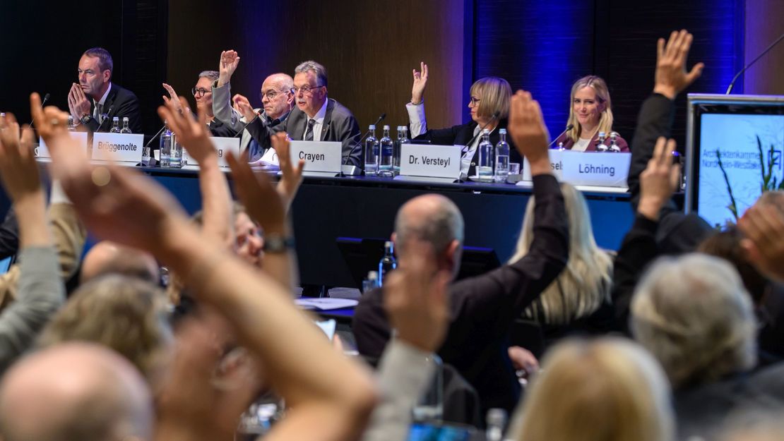 Zahlreiche Menschen sitzen in einem Raum, einige von ihnen heben die Hand zu einem Handzeichen.