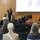 Ein bestuhlter Raum, viele Personen sitzen. An einem Pult vorne im Raum steht ein Mann und zeigt auf einer Leinwand eine digitale Präsentation mit verschiedenen Grafiken.