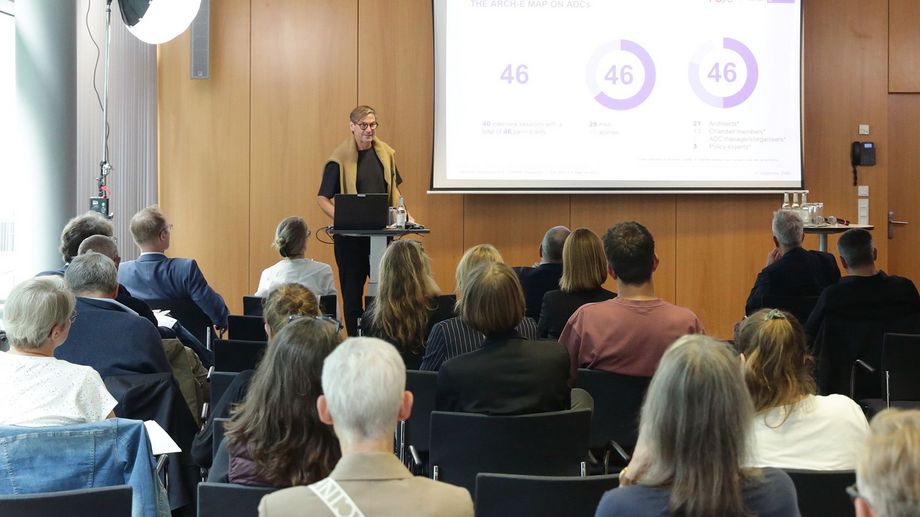 Ein bestuhlter Raum, viele Personen sitzen. An einem Pult vorne im Raum steht ein Mann und zeigt auf einer Leinwand eine digitale Präsentation mit verschiedenen Grafiken.