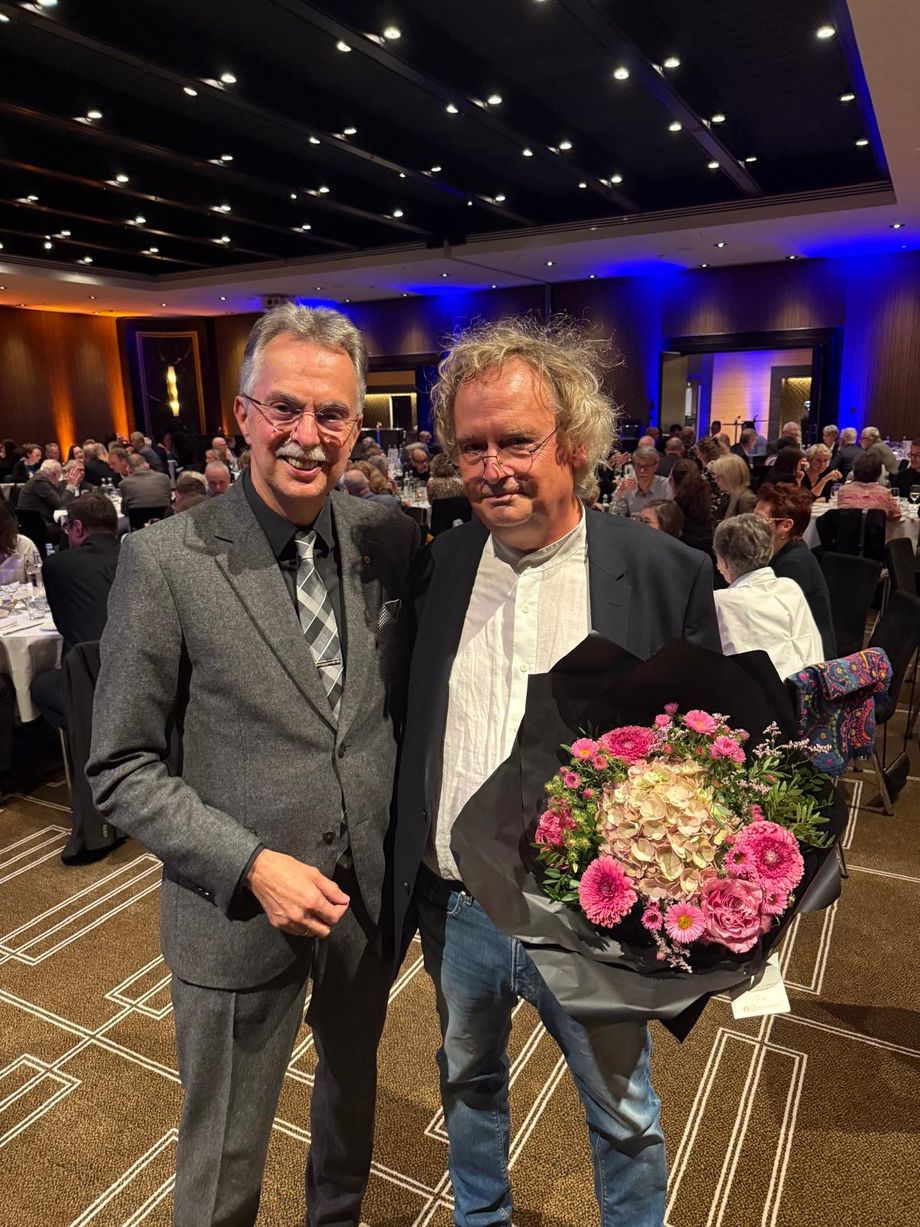 Zwei Männer stehen in einem Raum und lächeln in die Kamera, ein Mann hält einen bunten Blumenstrauß fest.