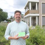 Ein Mann hält einen Katalog zum "Tag der Architektur 2025" fest und lächelt in die Kamera. Im Hintergrund ist ein Haus zu sehen und grüne Bepflanzung. 