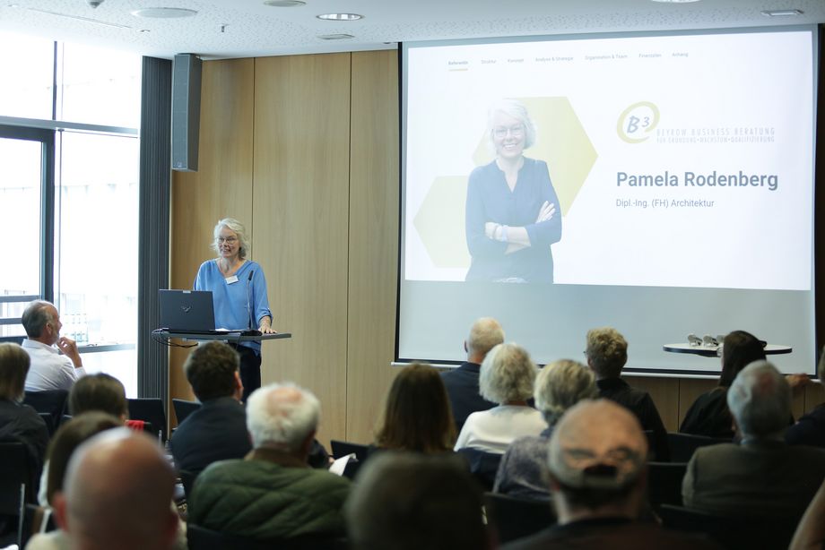 Eine Frau steht an einem Pult vor einer Leinwand, auf der eine Präsentation gezeigt wird. Im Vordergrund sitzen Leute und hören der Frau zu. - Foto: Ingo Lammert/Architektenkammer NRW