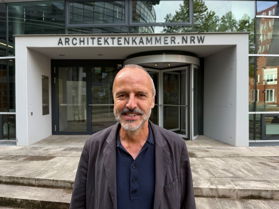 Ein Mann steht vor einem Gebäude und lächelt. Über dem Eingang des Gebäudes steht die Aufschrift "ARCHITEKTENKAMMER.NRW". - Foto: Christof Rose/Architektenkammer NRW