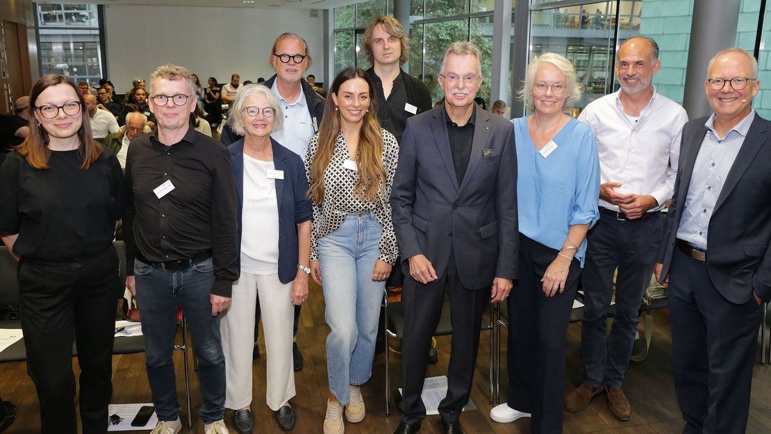 AKNW-Existenzgründungstag - Gruppenbild – Foto: Ingo Lammert/Architektenkammer NRW Eine Gruppe von Männern und Frauen, die lächelnd in die Kamera blicken. Im Hintergrund sitzen Teilnehmende einer Veranstaltung auf Stühlen. – Foto: Ingo Lammert/Architektenkammer NRW