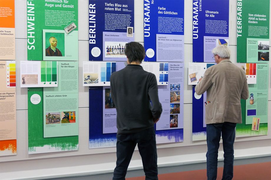 Zwei Männer stehen vor bunten Informationstafeln zum Thema Farben und Pigmente und schauen sich die Informationstafeln an.