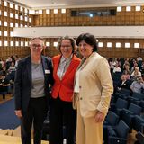 Drei Personen stehen auf einer Bühne beieinander, im Hintergrund sitzen Leute im Publikum.