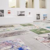 Blick in einen hellen Ausstellungsraum, auf weißen Sockeln stehen Fotografien gerahmt in durchsichtigen Kästen, auf dem Boden liegen bunte Teppiche, die Fotografien und Bilder abbilden.