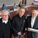 Drei Männer stehen lächeln und stehen beeinander, im Hintergrund sitzen mehrere Personen an einem Tisch.