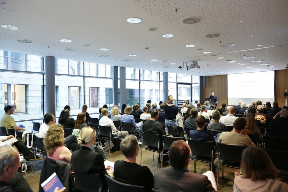 Ein bestuhlter Raum mit sitzenden Personen. Im Vordergrund steht ein Mann an einem Pult und zeigt eine Präsentation auf einer Leinwand