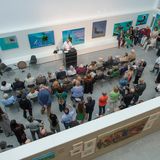 Menschen stehen und sitzen in einem hellen Ausstellungsraum. An einem Pult steht eine Person und spricht. An den Wänden hängen bunte Gemälde.