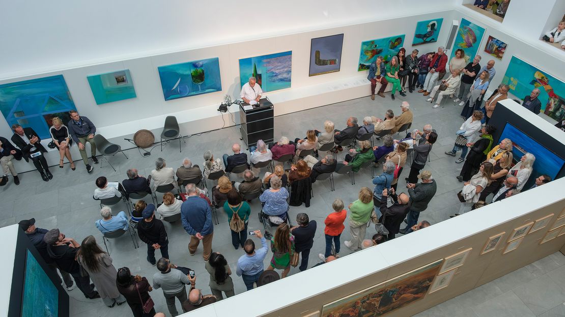 Menschen stehen und sitzen in einem hellen Ausstellungsraum. An einem Pult steht eine Person und spricht. An den Wänden hängen bunte Gemälde.
