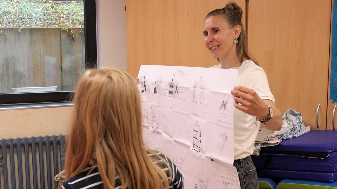 Eine Person hält einen Bauplan fest und präsentiert diesen. Im Vordergrund steht eine Person und betrachtet den Plan.