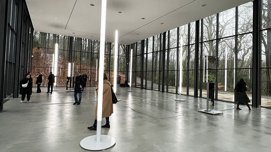 Ausstellungshalle mit großen Glasfronten, durch die man auf den Wald blickt, mit der aus vielen Lichtstelen bestehenden Installation
