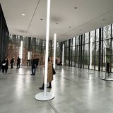 Ausstellungshalle mit großen Glasfronten, durch die man auf den Wald blickt, mit der aus vielen Lichtstelen bestehenden Installation