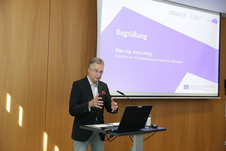 Ein Mann steht an einem Pult und spricht in ein Standmikrofon. Im Hintergrund ist eine Leinwand zu sehen. Gezeigt wird eine Präsentation, die Folie zeigt die Beschriftung: "Begrüßung. Dipl.-Ing. Ernst Uhing. Präsident der Architektenkammer Nordrhein-Westfalen" 