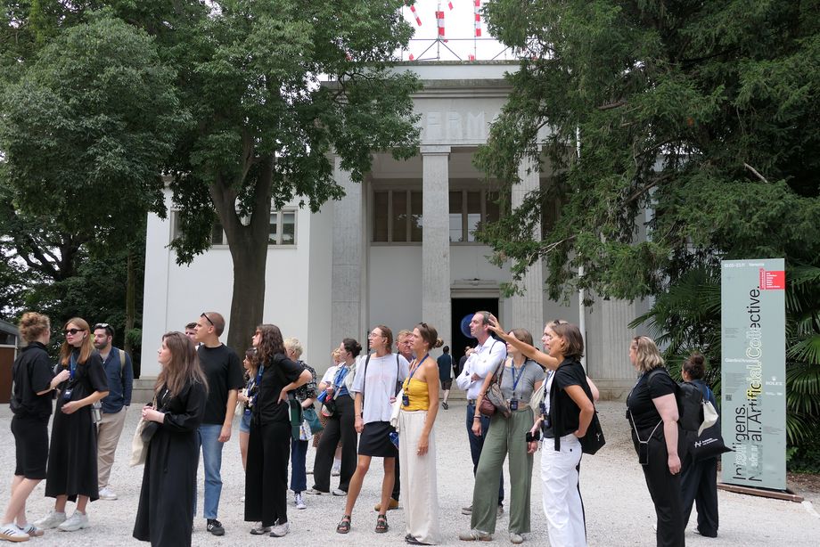 Eine Gruppe von Männern und Frauen steht vor einem hohen Gebäude mit hellen Säulen, vor dem auch einige Bäume stehen.