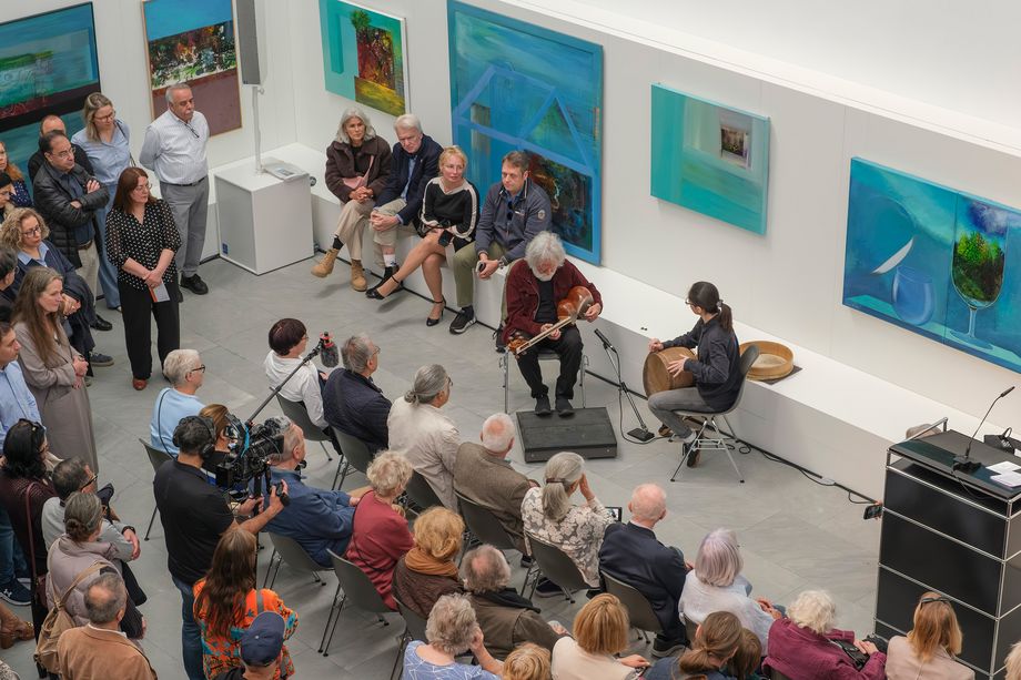 Ein heller Ausstellungsraum, an den Wänden sind bunte Gemälde. Personen sitzen und stehen. Einige Personen spielen Musikinstrumente.