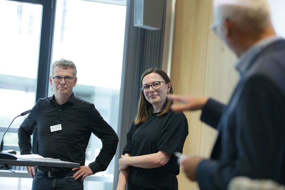 Eine Frau und ein Mann stehen an einem Pult. Im Vordergrund ist ein weiterer Mann zu sehen, der ein Gespräch mit den Personen am Pult führt. - Foto: Ingo Lammert/Architektenkammer NRW