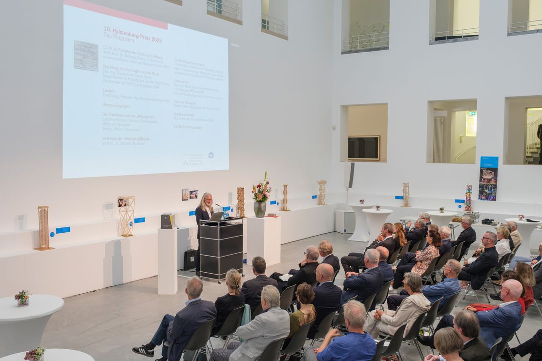 Halstenbergpreis 2025 - Foto: Detlef Podehl Eine Gruppe Menschen sitzt in einem großen Raum, an einem Pult steht ein Mann und hält einen Vortrag.