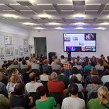 Eine Gruppe von Menschen in einem Raum, die teilweise sitzen und teilweise stehen. Eine Person steht mit einem Mikrofon in der Hand vor einer Leindwand.